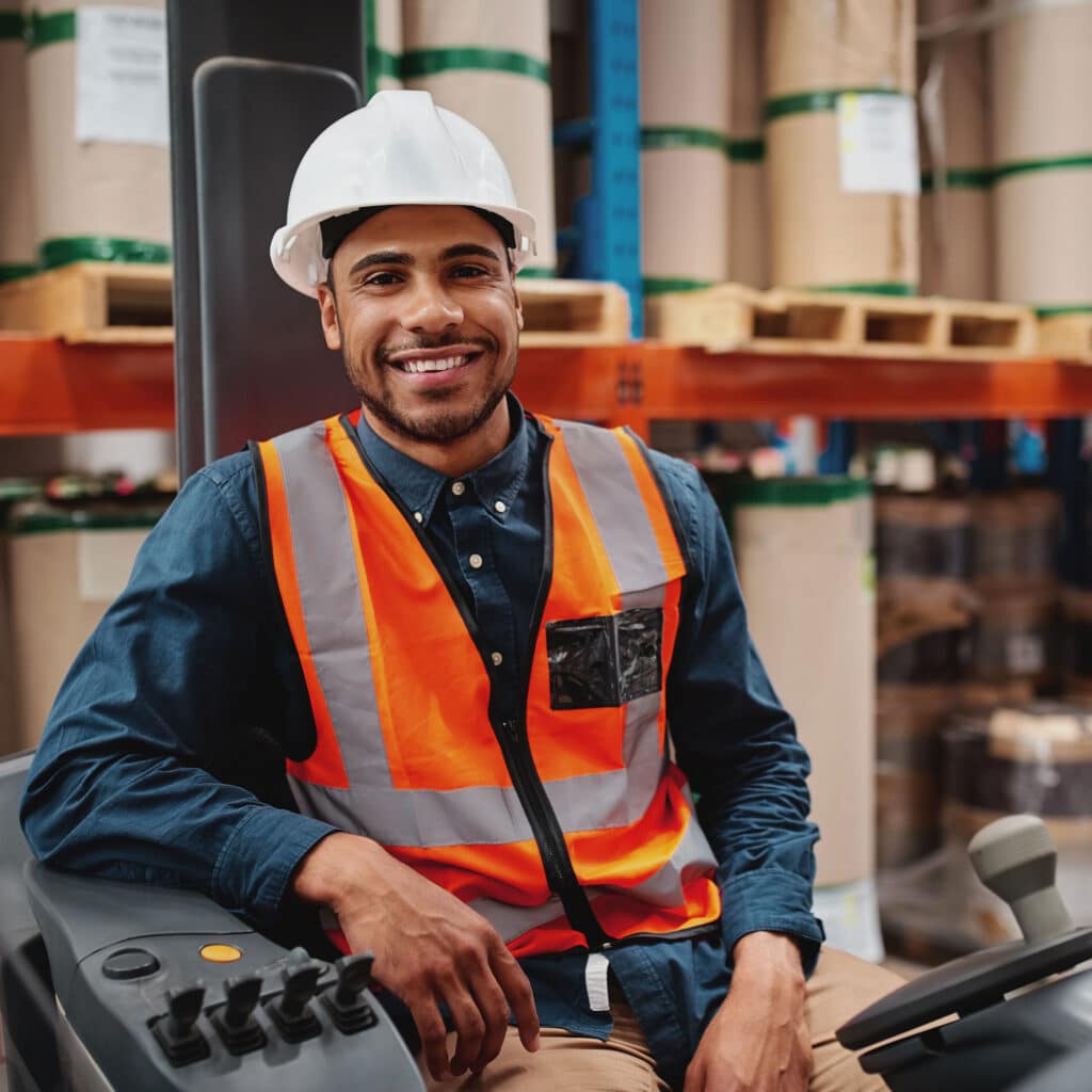 Handsome engineer wearing protective uniform and hardhat smiling joyfully to the camera sitting in forklift stacker while working at the storage
