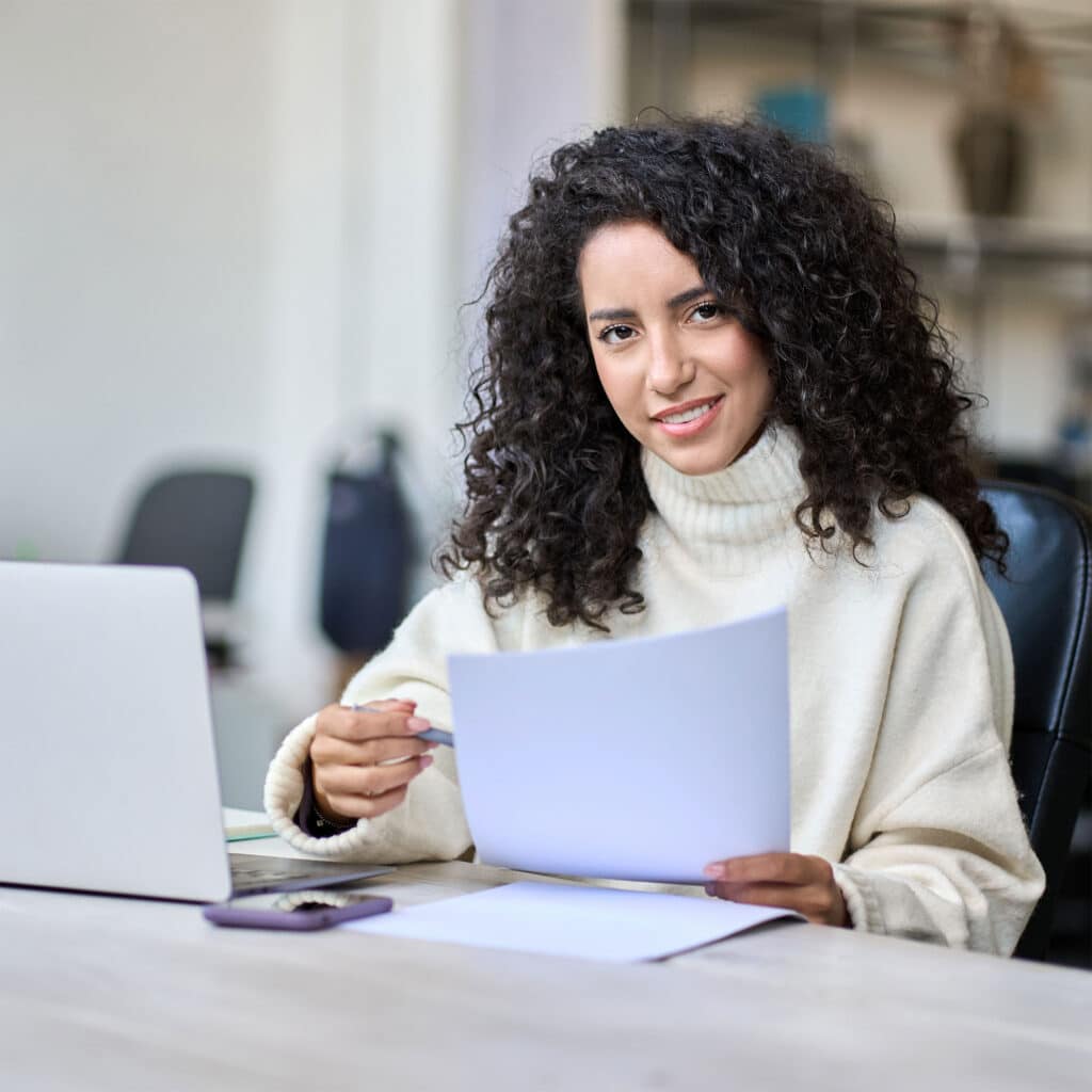 Young smiling latin business woman manager accounting analyst checking bills, analyzing sales management report, taxes financial data documents or marketing papers working in office using laptop.
